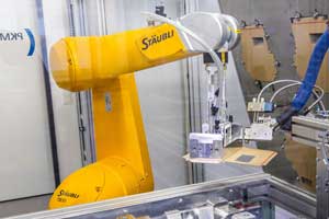 Per Roboterarm wird die benötigte Zuschnittgröße aus dem Vorrat entnommen, mit Heißkleber bestrichen und über eine Aufrichtschablone in den Transportwagen eingesetzt.